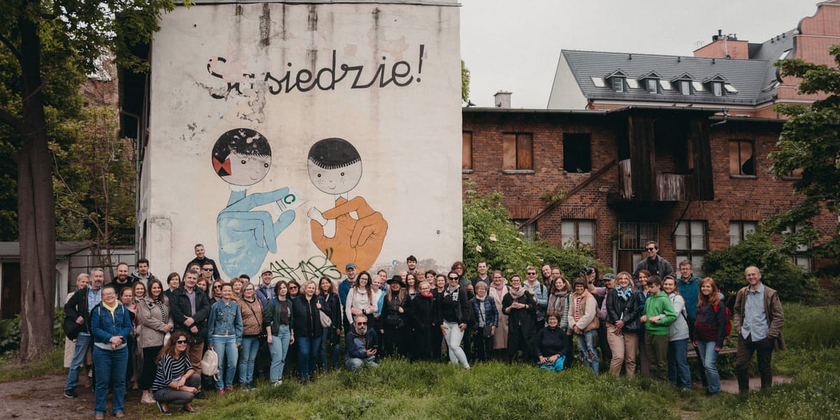 Uczestnicy spaceru sąsiedzkiego pozujący do grupowego zdjęcia w plenerze. W tle budynek z muralem. Zdjęcie archiwalne