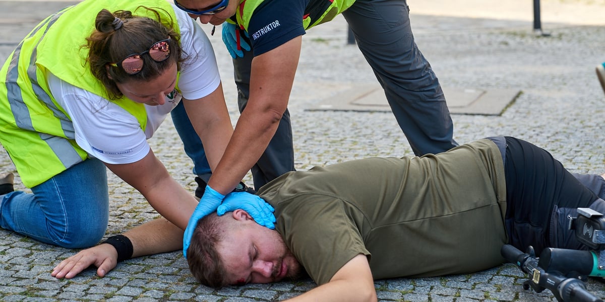 Na bruku leży mężczyzna w krótkich spodenkach, obok w odblaskowych kamizelkach klęczą dwie osoby i udzielają poszkodowanemu pierwszej pomocy