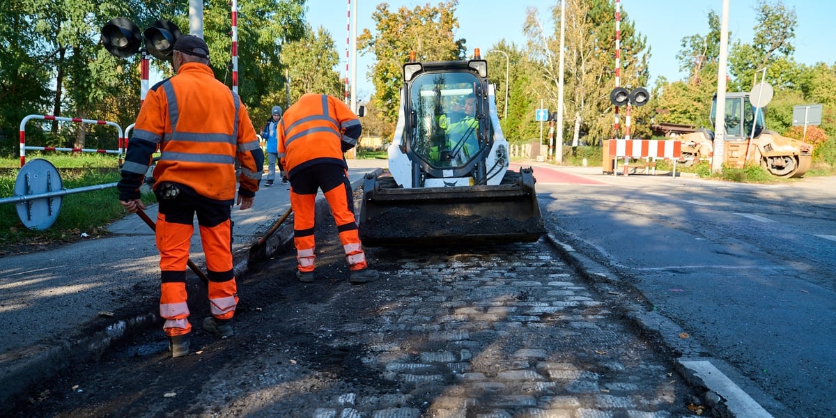 Dwaj mężczyźni w pomarańczowych kombinezonach pracują na ul. Ołtaszyńskiej, widać też maszynę budowalną 