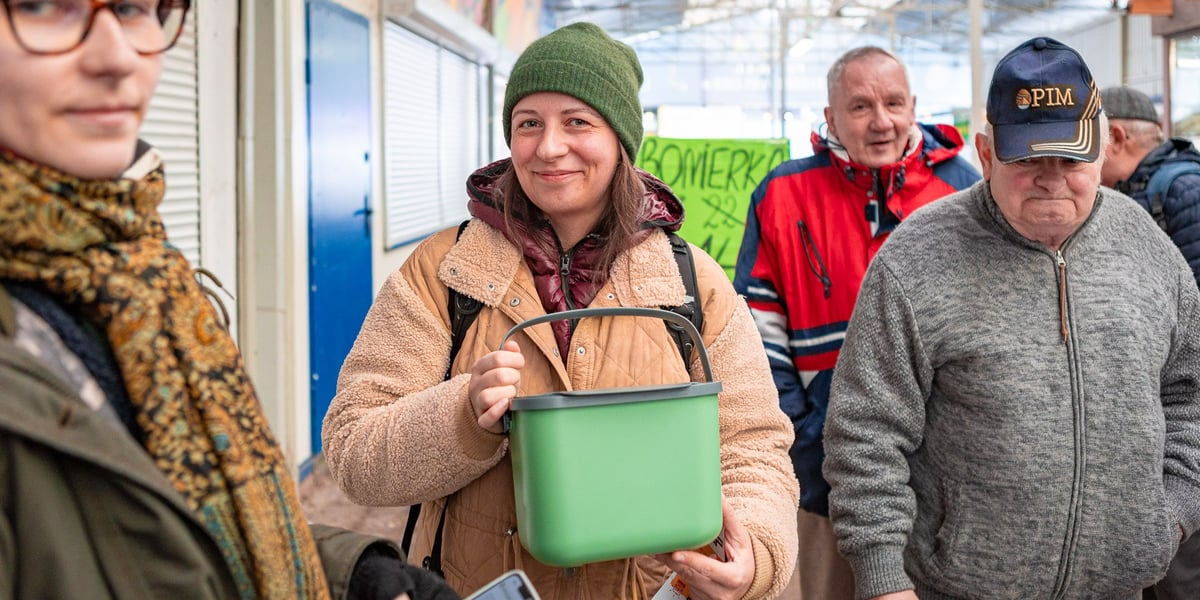 W centralnej części zdjęcia jest uśmiechnięta kobieta (w czapce i kurtce) trzymająca pojemnik na bioodpady na targowisku Komandor (7 grudnia 2024 r.)