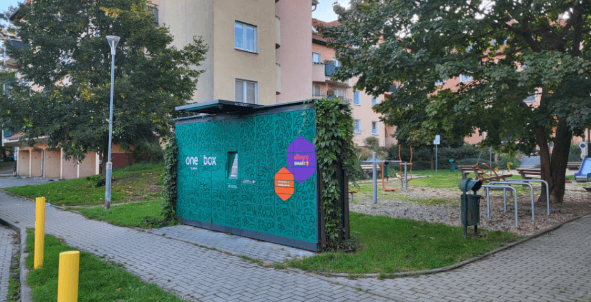 Automat kurierski oklejony zielonym motywem roślinnym, porośnięty rośliną pnącą, wzdłuż chodnika, na tle placu zabaw.