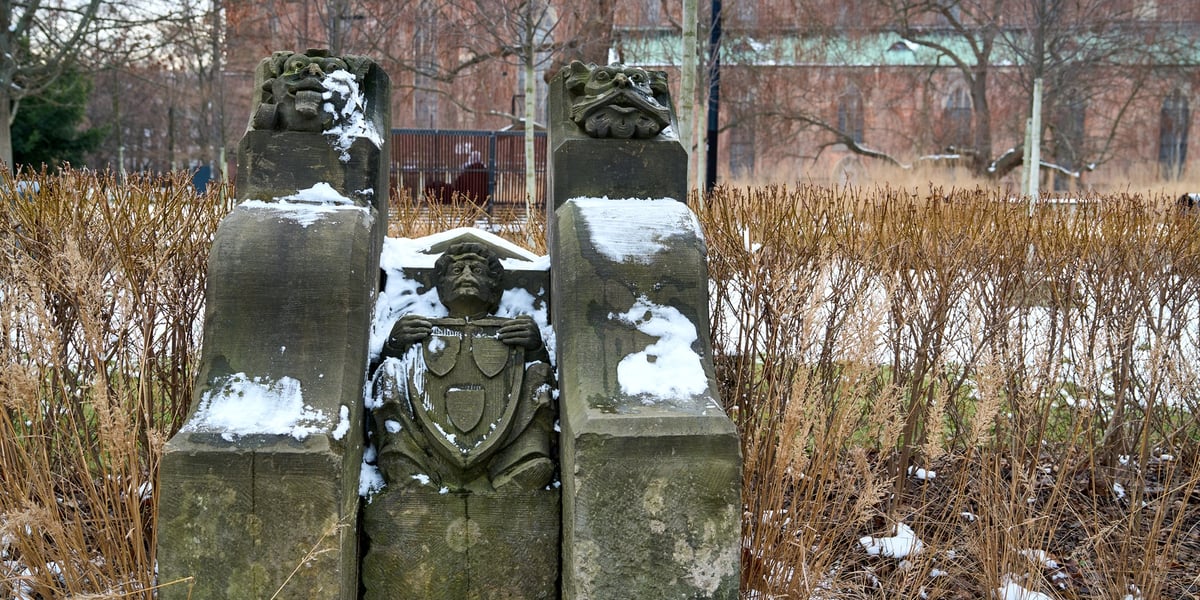 Fragment kamieniarki z rzeźbą mężczyzny i dwiema innymi przedstawiającymi zwierzęta, na tle placu i budynku z czerwonej cegły