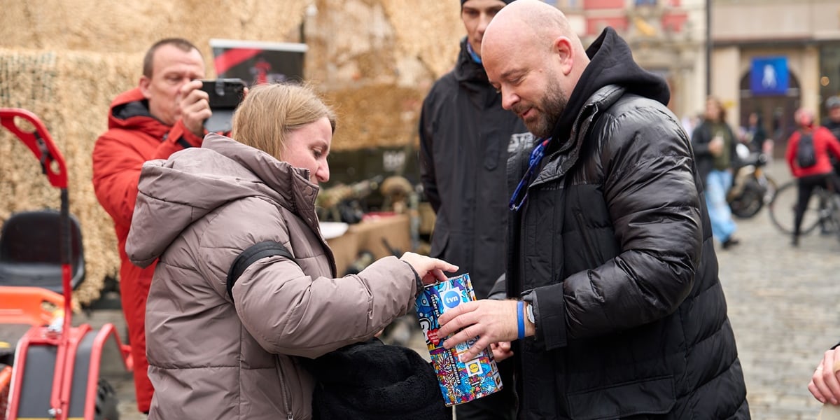 Prezydent Wrocławiu kwestuje podczas finału WOŚP. Na zdjęciu mężczyzna w płaszczu i kobieta wrzuca pieniądze do skarbonki WOŚP, którą trzyma prezydent Jacek Sutryk.