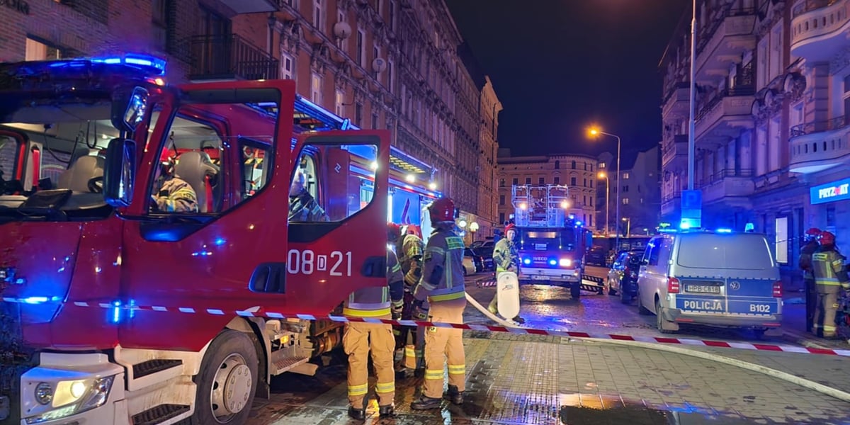 Wozy straży pożarnej, strażacy, niebieskie światła kogutów, kamienica, w tle ulica