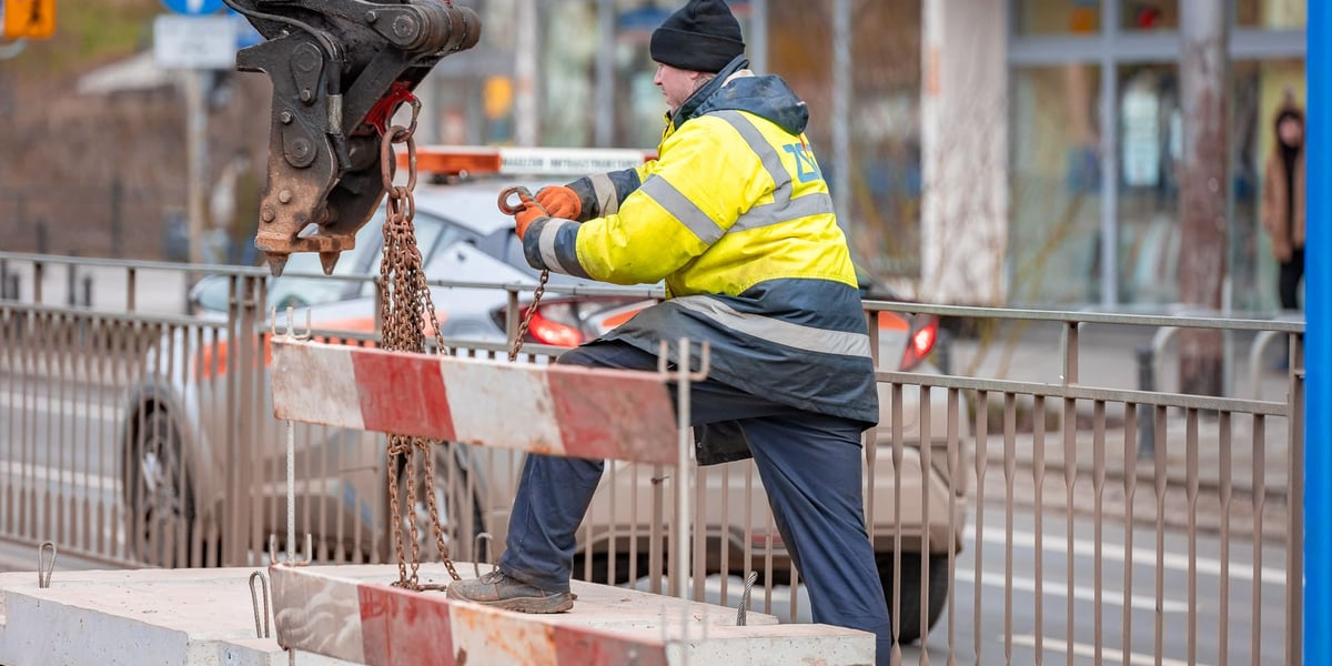 Remont torowiska na ul. Kołłątaja we Wrocławiu, 01.02.2025. Na zdjęciu mężczyzna w żółtej roboczej kamizelce przyczepia betonową płytę do ramienia koparko-ładowarki. 