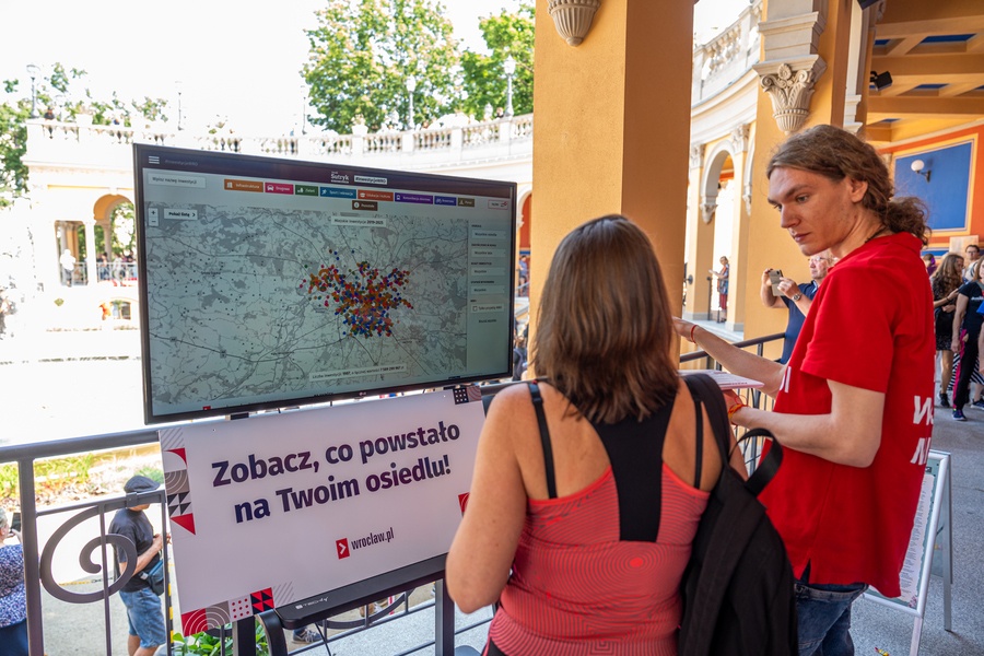 Kobieta i mężczyzna stoją przed monitorem, na którym wyświetlona jest baza #InwestycjeWRO