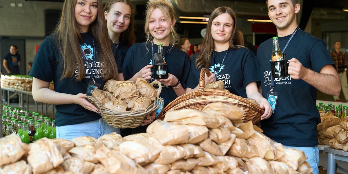 Rozpoczęcie Juwenaliów 2025 na Politechnice Wrocławskiej: śniadanie z rektorem. Na zdjęciu grupa młodych osób w koszulkach politechniki stoi przed stosem kanapek zapakowanych w papierowe torebki