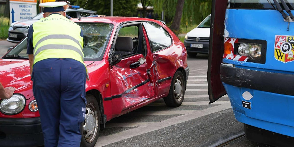 Czerwony samochód z pogniecionymi drzwiami po zderzeniu ze stojącym obok tramwajem, pracownicy nadzoru ruchu