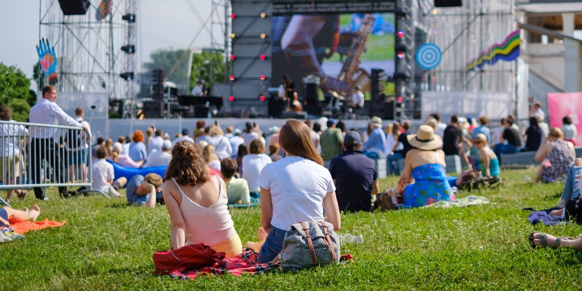 Koncert plenerowy. Zdjęcie ilustracyjne. Na zdjęciu widać ludzi, którzy siedzą na trawie przed duża sceną
