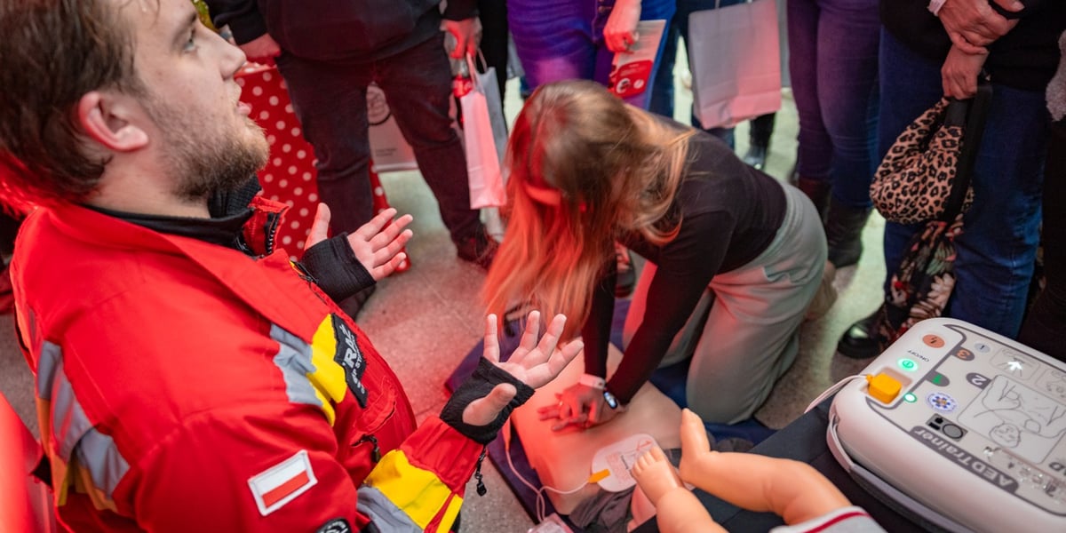Mężczyzna i kobieta podczas ćwiczeń z pierwszej pomocy, w tle stoją ludzie