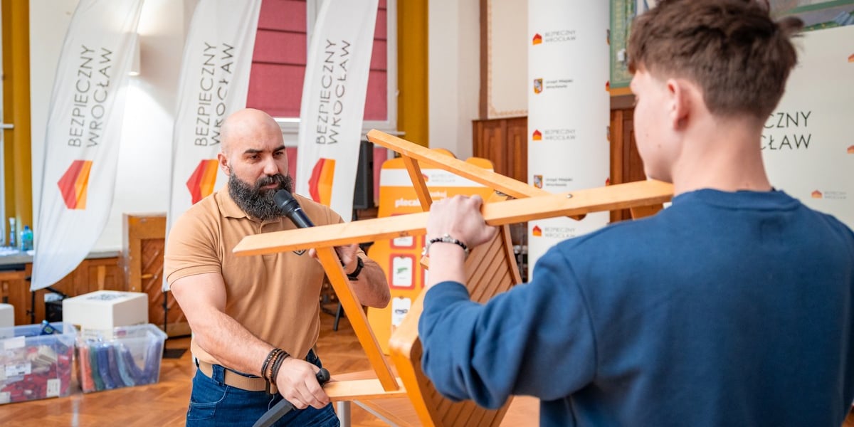 Szkolenie w ramach programu "Bezpieczny Wrocław" w I LO we Wrocławiu. Na zdjęciu mężczyzna z atrapą nożna i chłopak zasłaniający się krzesłem
