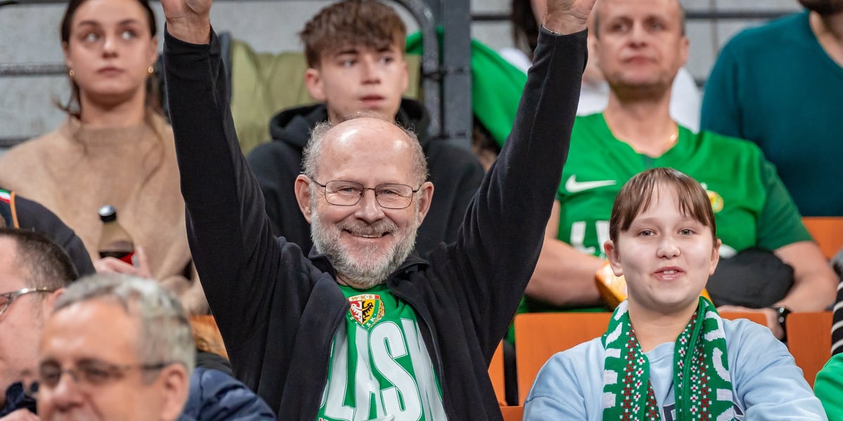 Uśmiechnięty mężczyzna w zielonej koszulce Śląska i bluzie, obok niego kobieta i inni kibice na trybunie Hali Stulecia na meczu z Arisem Saloniki, 20 stycznia 2026. 