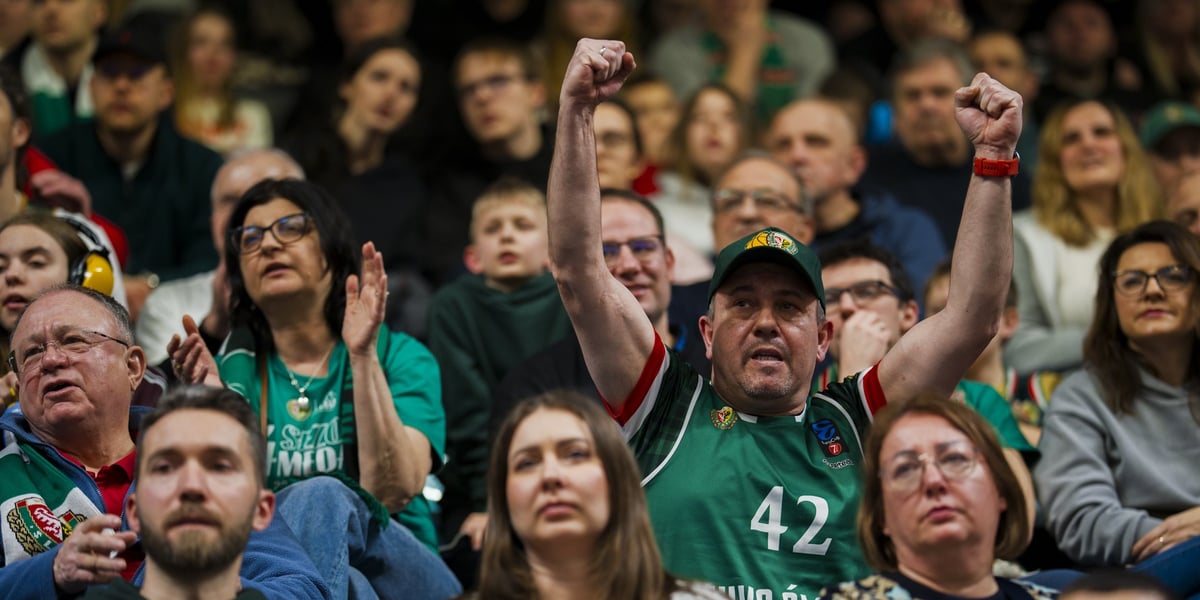 Kibic w zielonej koszulce Śląska i w czapce z daszkiem, unoszący ręce na trybunie Hali Orbita podczas spotkania z Gliwicami. Na zdjęciu są także inni kibice.