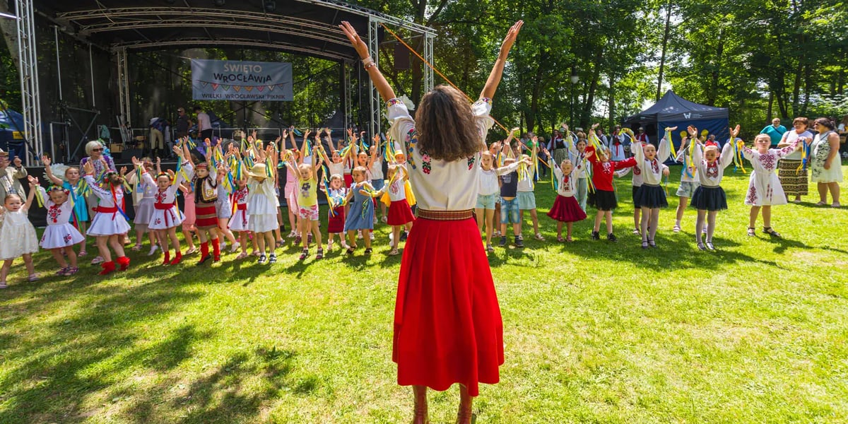 Przed grupą tańczących dzieci stoi kobieta w ludowym stroju i wyciąga ręce do góry