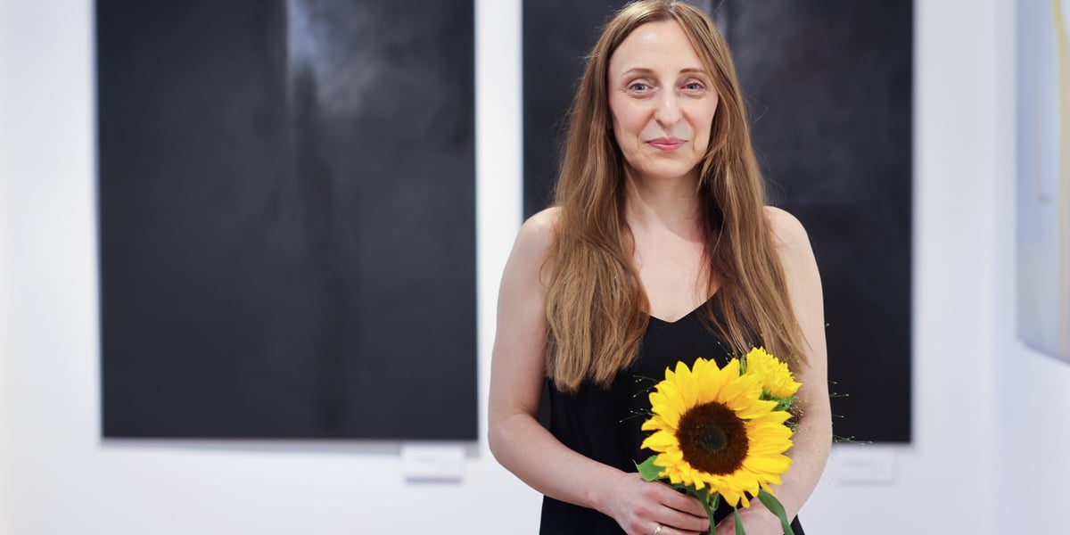 Agnieszka Semaniszyn-Konat ze słonecznikiem na tle swoich obrazów w Galerii Tkackiej na Jatkach, lipiec 2026
