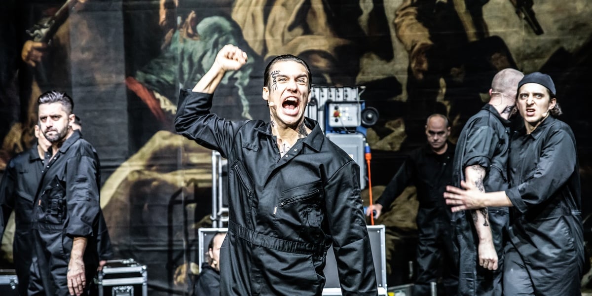 Na zdjęciu scena ze spektaklu "Marat/Sade" we Wrocławskim Teatrze Współczesnym. Na pierwszym planie ubrany na czarno mężczyzna z uniesioną do góry pięścią.