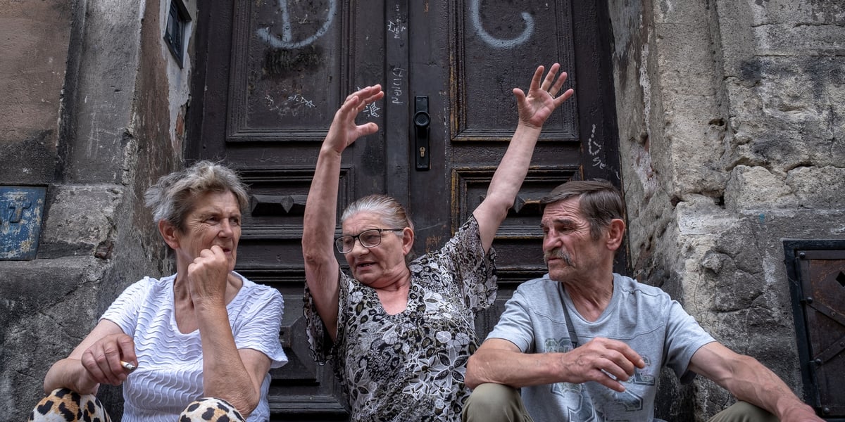 Diw kobiety i mężczyzna siedzą przed popisanymi drzwiami kamienicy w Legnicy, jedna z kobiet unosi ręce w ekspresyjnym geście