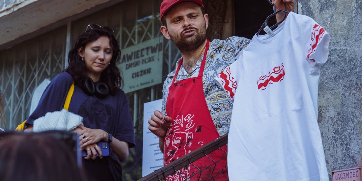 Jan Kowalski w fartuchu pokazuje koszulkę z biało-czerwoną flagą przed sklepem w krawiectwie ciężkim 