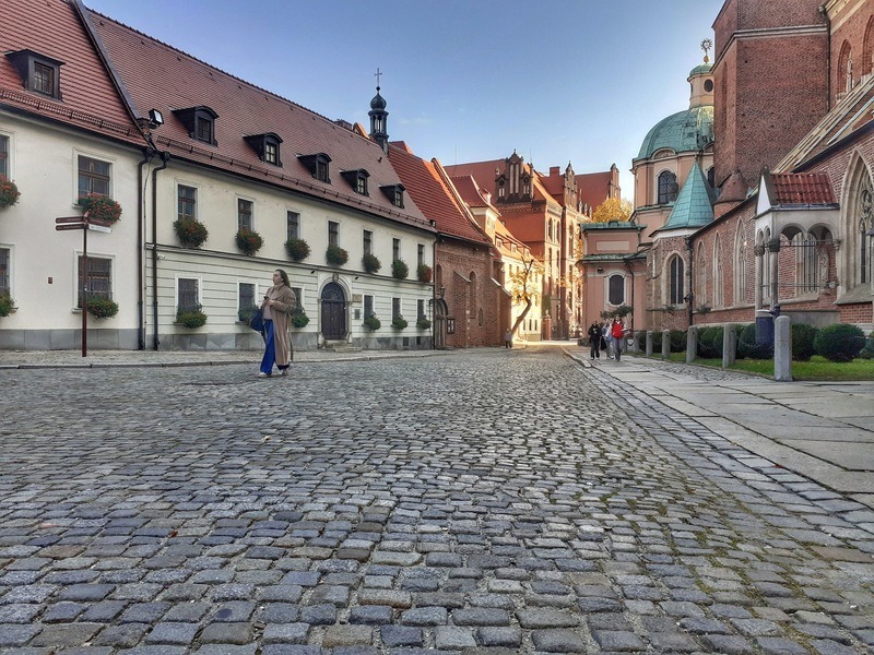 Na pierwszym planie jest kostka brukowa Placu Katedralnego (spaceruje po nim kobieta), w XIV wieku był tu cmentarz parafii diecezjalnej na Wyspie Tumskiej