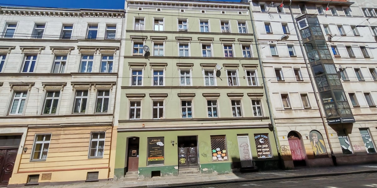 Zielona kamienica z punktami usługowymi na parterze, stojąca między jasnymi budynkami przy ulicy Szczytnickiej z torami tramwajowymi. Zobacz, gdzie gmina Wrocław sprzedaje lokale użytkowe. 