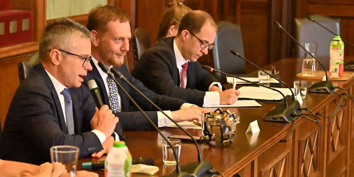 Trzech mężczyzn w garniturach siedzi przy podłużnym stole, przed nimi stoją mikrofony, leżą notatniki. Delegacja Saksonii w Ratuszu 