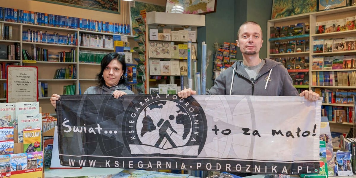 Pracownicy Księgarni Podróżnika trzymają baner z napisem "Świat to za mało.. Księgarnia Podróżnika", w tle regały z książkami i mapami