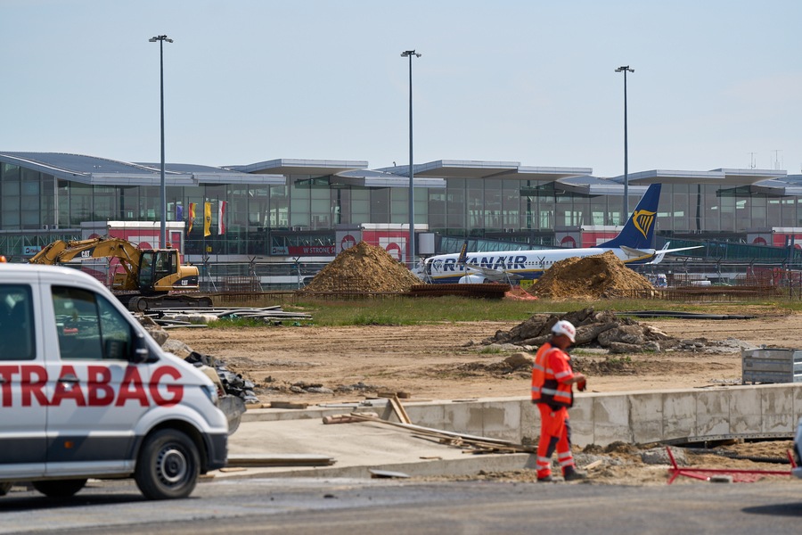 Na pierwszym planie jest plac budowy z robotnikiem, w głębi widać samolot i terminal wrocławskiego lotniska