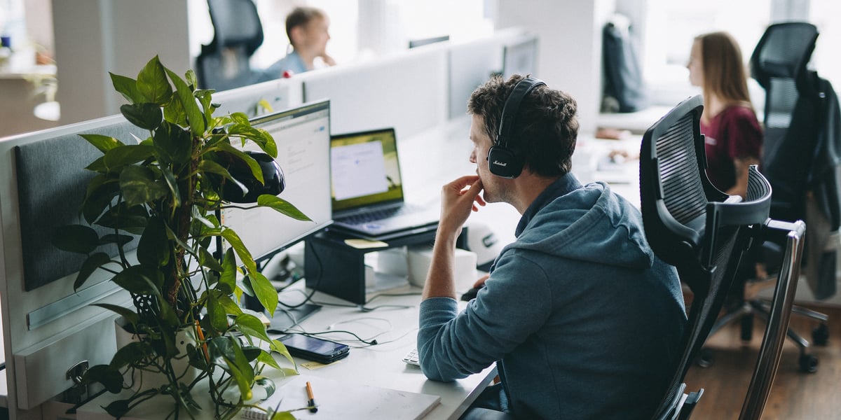 Mężczyzna w słuchawkach patrzy na monitory komputerów, w tle kobieta i monitory. Pracownicy spółki Monterail