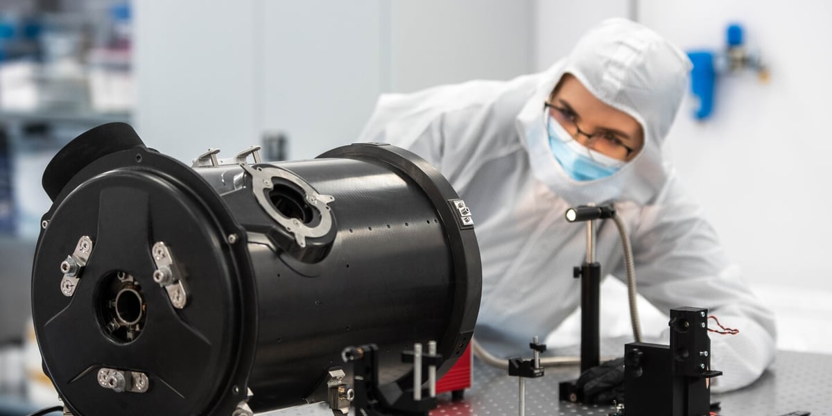 Elementy konstrukcyjne teleskopu, w tle kobieta w okularach w kombinezonie i z maseczką na ustach w laboratorium firmy Scanway. Wrocławski spółka buduje największy w Polsce teleskop optyczny.