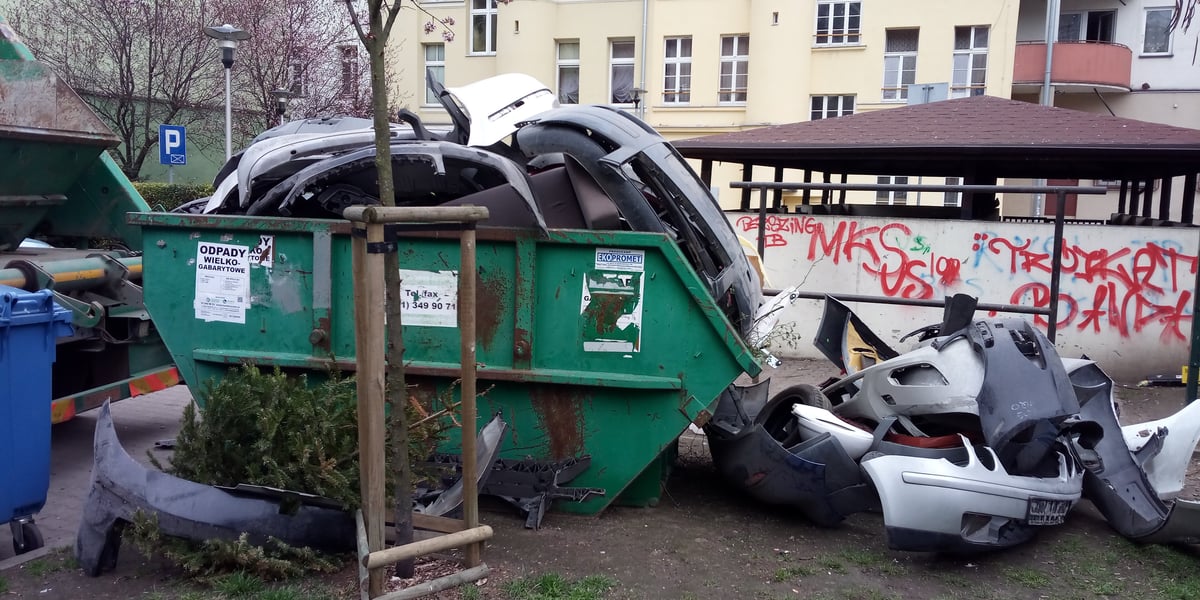 Zielony kontener na odpady wielkogabarytowe wypełniony częściami samochodu, które leżą także obok