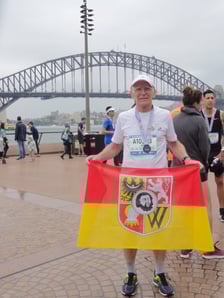 Biegacz na zdjęciu w stroju sportowym to prof. Jan Chmura. Trzyma czerwono-żółtą flagę z herbem Wrocławia, którą zawsze zabiera ze sobą na maratony. Zdjęcie zrobione po zakończeniu Blackmores Sydeny Marathon na tle słynnego Harbour Bridge. Wrocławianin startował w ramach Korony 7 Kontynentów. Zajął 3. miejsce w kategorii 65-69