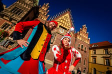 Dwoje szczudlarzy w barwnych, bajkowych strojach krasnali pozuje na tle zabytkowego Starego Ratusza we Wrocławiu