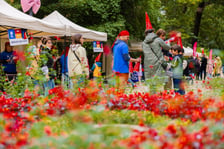 Ludzie odwiedzają plenerowe stoiska Festiwalu Krasnali w parku, widoczni zza rozmytego planu pełnego czerwonych kwiatów