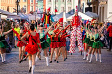 Grupa młodych tancerek w strojach estradowych maszeruje w radosnej paradzie na wrocławskim Rynku w towarzystwie szczudlarzy przebranych za krasnale