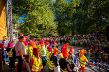 Liczna grupa dzieci w odblaskowych kamizelkach i pomarańczowych czapkach krasnali gromadzi się w parku przy plenerowej scenie podczas Festiwalu Krasnali