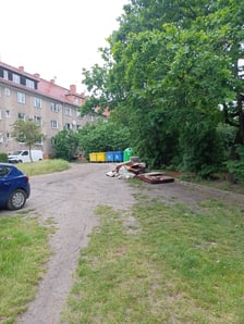 Na podwórzu widać wyjeżdżoną przez auta trawę i dwa parkujące na niej samochody. W głębi stoją śmietniki, przed nimi, na ziemi, leżą duże odpady.