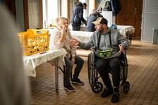 <p>mężczyzna na w&oacute;zku inwalidzkim oraz siedząca na krześle kobieta, obok st&oacute;ł nakryty ceratą, na kt&oacute;rym stoi skrzynka z pieczywem</p>