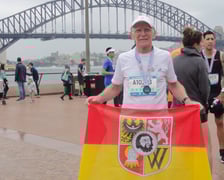 Biegacz na zdjęciu w stroju sportowym to prof. Jan Chmura. Trzyma czerwono-żółtą flagę z herbem Wrocławia, którą zawsze zabiera ze sobą na maratony. Zdjęcie zrobione po zakończeniu Blackmores Sydeny Marathon na tle słynnego Harbour Bridge. Wrocławianin startował w ramach Korony 7 Kontynentów. Zajął 3. miejsce w kategorii 65-69