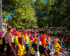 Liczna grupa dzieci w odblaskowych kamizelkach i pomarańczowych czapkach krasnali gromadzi się w parku przy plenerowej scenie podczas Festiwalu Krasnali