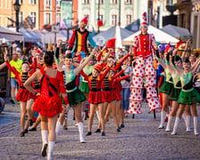 Grupa młodych tancerek w strojach estradowych maszeruje w radosnej paradzie na wrocławskim Rynku w towarzystwie szczudlarzy przebranych za krasnale