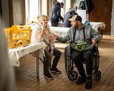 <p>mężczyzna na w&oacute;zku inwalidzkim oraz siedząca na krześle kobieta, obok st&oacute;ł nakryty ceratą, na kt&oacute;rym stoi skrzynka z pieczywem</p>