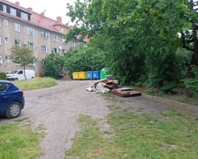 Na podwórzu widać wyjeżdżoną przez auta trawę i dwa parkujące na niej samochody. W głębi stoją śmietniki, przed nimi, na ziemi, leżą duże odpady.