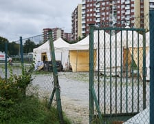 Chodnik osiedlowy. Przy nim ogrodzony teren placu targowego. Otwarta furtka - wejście na teren targowiska. Stanowiska handlowe w postaci białych namiotów. W tle zabudowania osiedla i duże bloki.