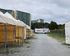 Teren targowiska. Biała, żwirowa nawierzchnia. Po lewej i prawej stronie stanowiska handlowe - białe namioty. W tle biały samochód dostawczy.