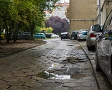 Zaniedbana, dziurawa, podwórkowa ulicę z betonowych płyt. Po prawej przy budynku parkujące w samochody. Po lewej skwer i parkujące wśród drzew samochody.