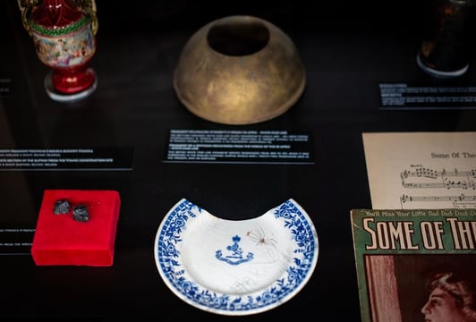 Zbliżenie na gablotę muzealną z artefaktami z liniowców, w tym fragmentem porcelanowego talerza w biało‑niebieski wzór, czerwonym pudełkiem z dwoma małymi obiektami oraz metalową misą.