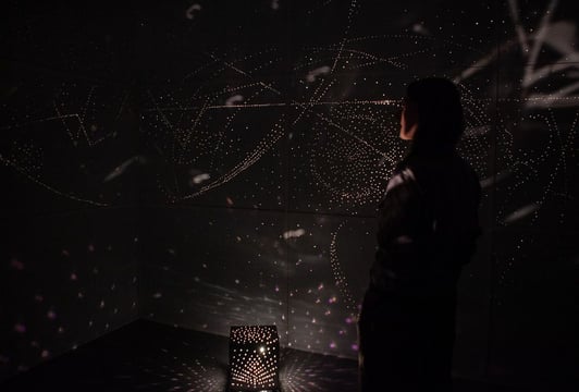 Otto Piene "Praska przestrzeń światła", fot. Magdalena Lorek. Ciemna przestrzeń w której na ścianach widać jasne świetliste punkty wydzielane przez małe świecące urządzenie ustawione na podłodze.
