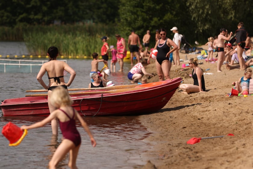 Kąpielisko Glinianki. Grupa os&oacute;b na plaży w strojach kąpielowych, widać też czerwoną ł&oacute;dź, dziecko z czerwonym wiaderkiem weszło do wody