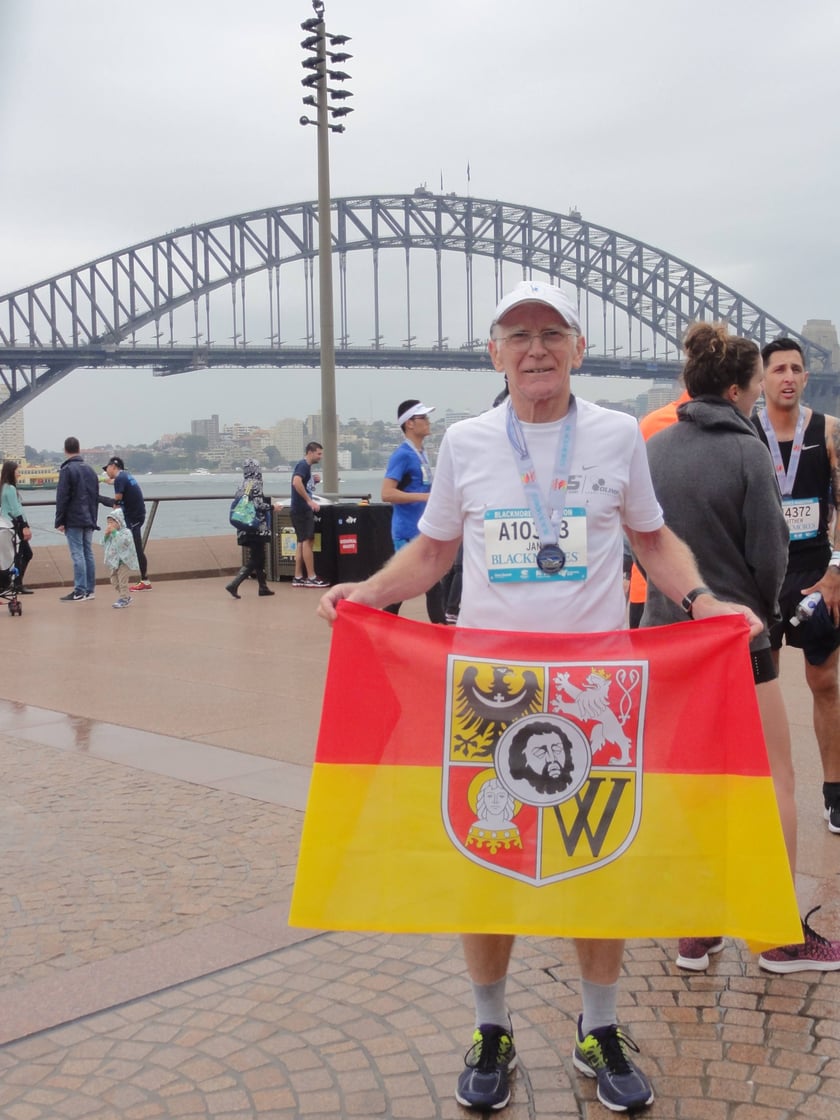 Biegacz na zdjęciu w stroju sportowym to prof. Jan Chmura. Trzyma czerwono-żółtą flagę z herbem Wrocławia, którą zawsze zabiera ze sobą na maratony. Zdjęcie zrobione po zakończeniu Blackmores Sydeny Marathon na tle słynnego Harbour Bridge. Wrocławianin startował w ramach Korony 7 Kontynentów. Zajął 3. miejsce w kategorii 65-69