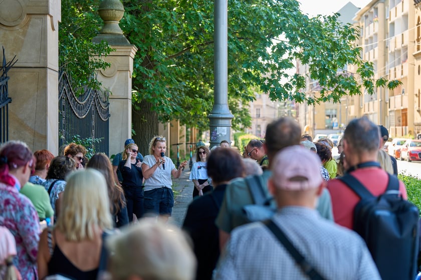 Przewodniczka z mikrofonem przemawia do grupy kilkudziesięciu uczestników spaceru stojących na chodniku przy ozdobnej, kutej bramie zabytkowego budynku, w tle widoczna miejska ulica z drzewami.